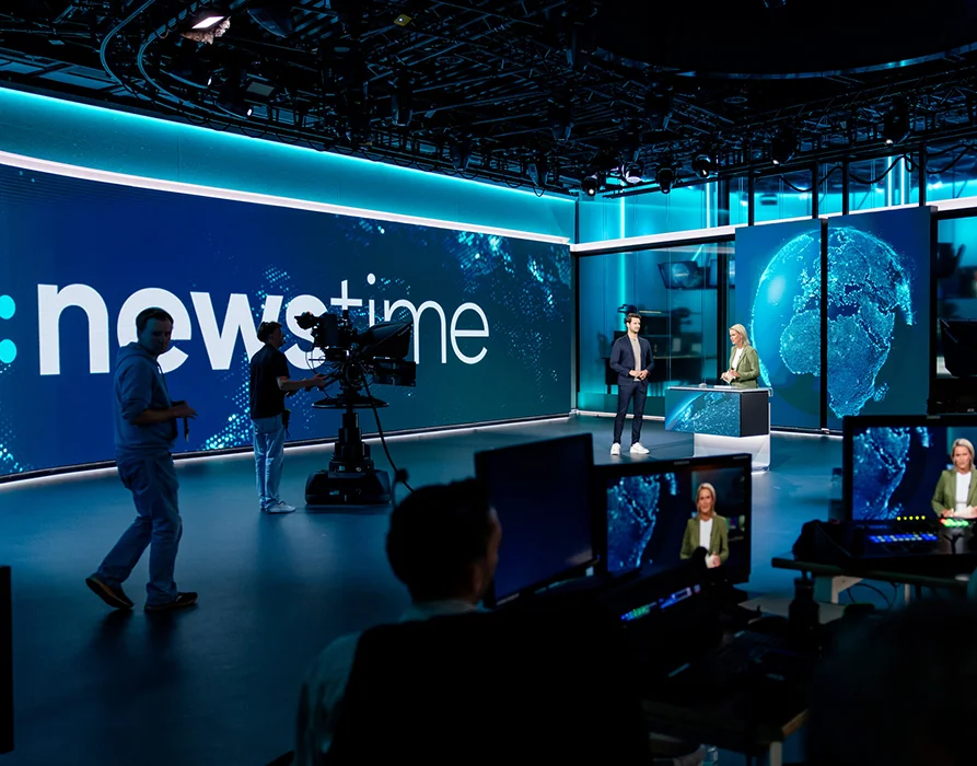 News studio with several screens and 4 people: someone at the camera, two presenters and one person walks by. The background shows the words ‘news time’ in large letters. (Photo)