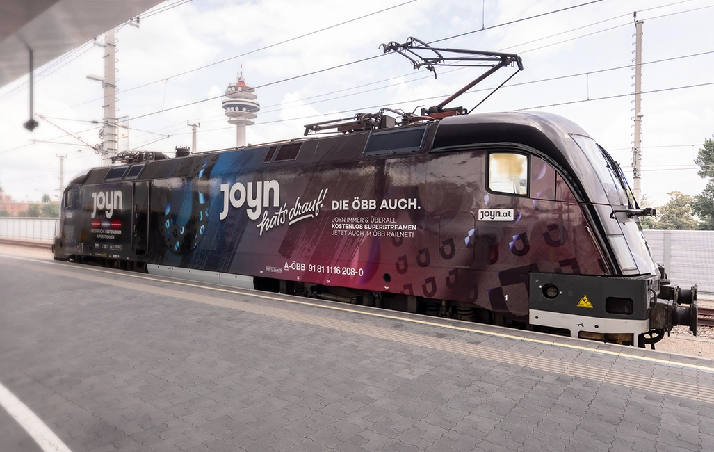 Railjet train engine of Austrian Federal Railways (known as ÖBB) covered with a Joyn ad and the slogan saying: "Joyn’s got it – so does ÖBB." The train engine is standing at the platform of a railway station. (Photo)